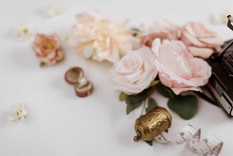 May include: A romantic still life featuring a velvet ring box with a diamond ring, blush-colored roses, and a vintage gold bell. A measuring tape and scattered flower petals complete the scene, evoking a sense of elegance and celebration.