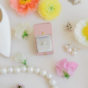 Puede incluir: Un anillo de compromiso de diamantes en una caja de terciopelo rosa, rodeado de perlas, flores y pendientes. La escena se desarrolla sobre una superficie blanca, con un zapato blanco en la esquina superior izquierda. Las flores son rosas, amarillas y blancas.