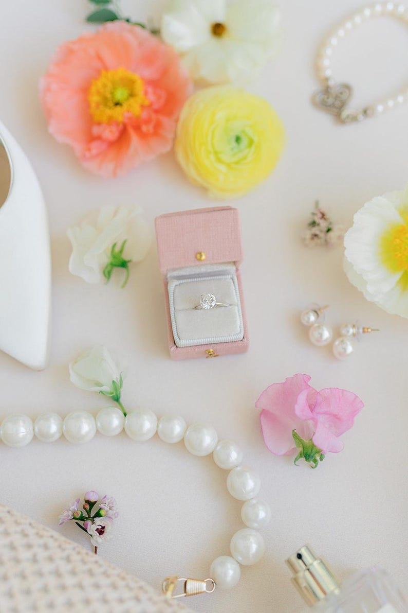 Puede incluir: Un anillo de compromiso de diamantes en una caja de terciopelo rosa, rodeado de perlas, flores y pendientes. La escena se desarrolla sobre una superficie blanca, con un zapato blanco en la esquina superior izquierda. Las flores son rosas, amarillas y blancas.