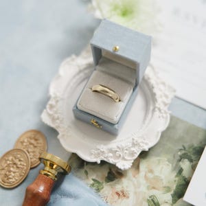 Puede incluir: Un anillo de bodas dorado descansa en un joyero azul claro con cierre dorado, sobre un plato decorativo blanco. La escena incluye un sello de cera, un sello de madera y oro, y papeler&iacute;a floral, sugiriendo un tema de boda.