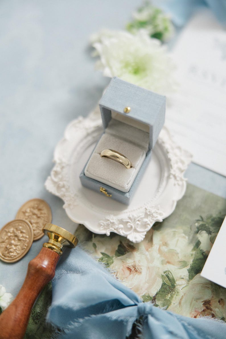 Puede incluir: Un anillo de bodas dorado descansa en un joyero azul claro con cierre dorado, sobre un plato decorativo blanco. La escena incluye un sello de cera, un sello de madera y oro, y papeler&iacute;a floral, sugiriendo un tema de boda.