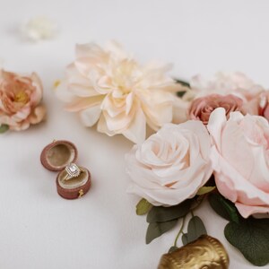 May include: A diamond engagement ring in a velvet ring box, surrounded by blush and cream-colored roses and other flowers. The ring has a square-cut diamond with a halo setting. The flowers are arranged on a white surface.