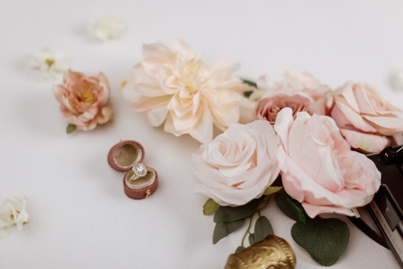 May include: A diamond engagement ring in a velvet ring box, surrounded by blush and cream-colored roses and other flowers. The ring has a square-cut diamond with a halo setting. The flowers are arranged on a white surface.