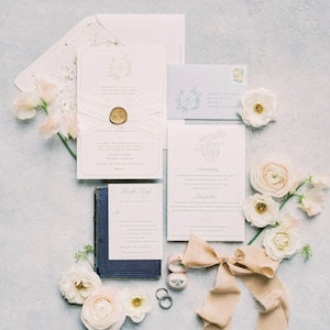 May include: A wedding stationery suite featuring a main invitation with a gold wax seal and white ribbon, a reply card resting on a dark blue book, an information card, a light blue envelope, two silver rings, a pink ring box, and beige silk ribbon.