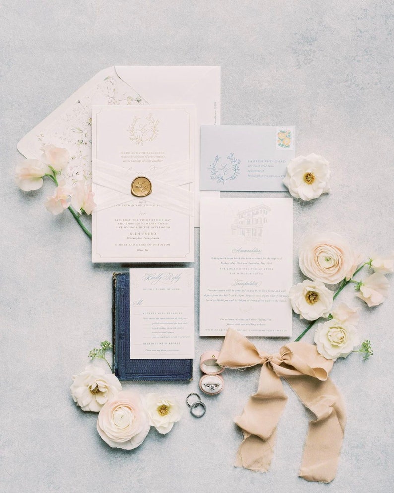 May include: A wedding stationery suite featuring a main invitation with a gold wax seal and white ribbon, a reply card resting on a dark blue book, an information card, a light blue envelope, two silver rings, a pink ring box, and beige silk ribbon.