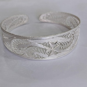 Puede incluir: Un brazalete de filigrana plateada con un diseño abierto. El brazalete presenta intrincados patrones florales y de estrellas. El diseño de brazalete abierto permite un fácil uso. El brazalete está hecho de un metal brillante y reflectante.