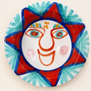 May include: A round ceramic bowl featuring a hand-painted sun face design. The center displays a white face with orange outlines, pink cheeks, and blue eyes. The border consists of alternating red triangular sun rays and teal fan-shaped patterns.