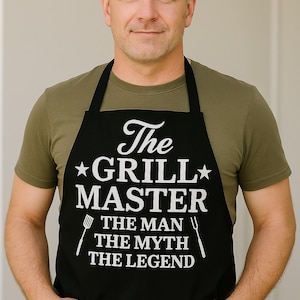 May include: A black apron with the text "The Grill Master" in white, along with "The Man, The Myth, The Legend". The apron is worn over a green t-shirt. The image features a person with their hands in their pockets.