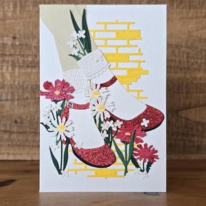 May include: A white card with a yellow brick road design. Red glitter shoes with white socks and white flowers are on the brick road.