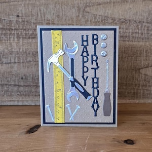 Może przedstawiać: Brązowa kartka z czarną ramką i brązowym tłem. Kartka ma motyw czarnych i srebrnych narzędzi z napisem "Happy Birthday" czarnymi literami. Kartka zawiera linijkę, młotek, klucz, śrubokręt i gwoździe.