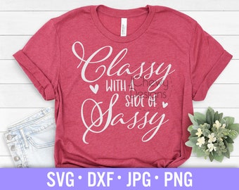 May include: A red t-shirt with the text "Classy with a side of Sassy" in white cursive font. The shirt is on a white wooden surface with a pair of blue jeans and a small potted plant.