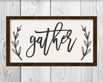 Op de afbeelding: Een wit bord met een bruin frame en het woord "gather" geschreven in zwarte cursieve letters. Het bord is versierd met twee zwarte, bladerrijke takjes aan weerszijden van het woord.