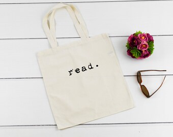 May include: A white canvas tote bag with the word "read." printed in black. The bag is on a white wooden surface with a small bouquet of pink flowers and a pair of sunglasses.