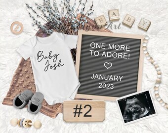 May include: A white onesie with the text "Baby Josh" in black script. A letter board with the text "One More To Adore! January 2023" and a heart symbol. A wooden block with the number "2" on it. A black and white ultrasound image.