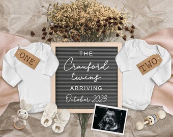 May include: A letter board with the text "The Crawford twins arriving October 2023". Two white baby onesies with the numbers "one" and "two" on them. A bouquet of dried flowers, a baby rattle, and a pair of white booties.