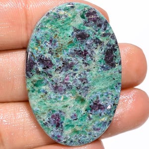 Puede incluir: Una piedra preciosa pulida de forma ovalada con un patrón moteado de inclusiones minerales en verde vibrante y morado intenso. La superficie muestra un acabado liso y brillante con texturas cristalinas visibles en toda la paleta de colores.