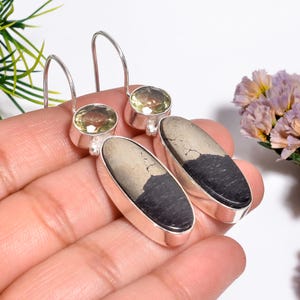 Può includere: Orecchini in argento con pietre di pirite ovali, nere e beige. Ogni orecchino ha una gemma rotonda, verde chiaro nella parte superiore. Gli orecchini sono tenuti in una mano, con una composizione floreale sullo sfondo.