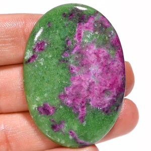 Puede incluir: Un cabujón de piedra preciosa pulida de forma ovalada que presenta una mezcla vibrante de zoisita verde intenso e inclusiones de rubí magenta brillante. La piedra tiene una superficie lisa y brillante con un patrón natural moteado.
