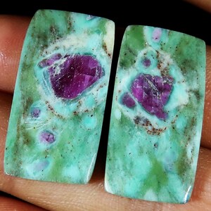 Könnte beinhalten: Zwei rechteckige Edelsteine mit einer lebendigen Farbpalette. Die Steine haben eine türkisfarbene Basis mit grüner und brauner Marmorierung. Jeder Stein hat einen zentralen, facettierten, tiefmagentafarbenen Einschluss.