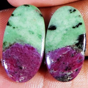 May include: Two oval-shaped gemstones. Each stone features a light green upper section with black inclusions, transitioning to a deep magenta lower section. The stones are polished, showcasing a glossy surface and natural patterns. These gemstones are suitable for jewelry making.