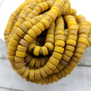 Pode incluir: Um fio de contas de vidro amarelo com uma textura ligeiramente áspera. As contas são redondas e têm um furo no centro. As contas são enfiadas em um cordão.