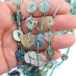 May include: A strand of antique glass beads, featuring a variety of colors and textures, including green, blue, and clear. The beads are strung on a thin green thread.