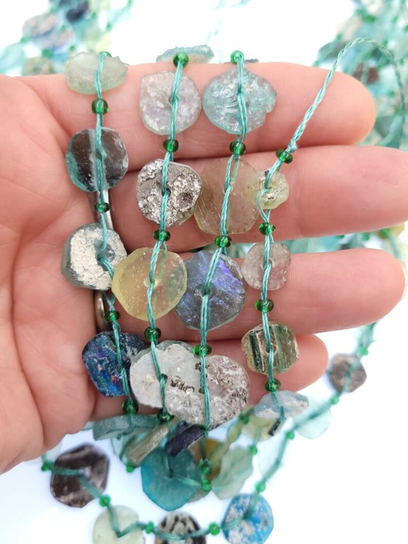 May include: A strand of antique glass beads, featuring a variety of colors and textures, including green, blue, and clear. The beads are strung on a thin green thread.