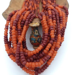 May include: A collection of beaded necklaces in shades of orange and burgundy, draped over a small, ornate vase. The beads are round and appear to be made of glass or a similar material. The vase has a detailed, colorful design.
