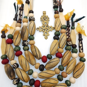 May include: A handcrafted necklace featuring a variety of wooden beads in natural tones, including oval, round, and cylindrical shapes. Accented with red, green, and black beads, and a gold-colored pendant. The necklace is adorned with fabric and feather accents.