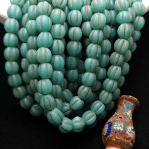 May include: A strand of turquoise blue glass beads with white stripes. The beads are round and have a slightly textured surface. A small, colorful, mosaic-style glass vase pendant hangs from the end of the strand.
