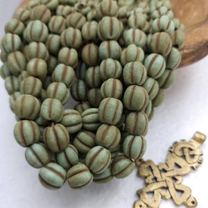 Pode incluir: Um fio de contas de vidro verde com listras marrons. As contas são redondas e têm uma textura ligeiramente irregular. As contas estão dispostas em uma pilha, e há um pequeno amuleto de cruz dourada na parte inferior da imagem.