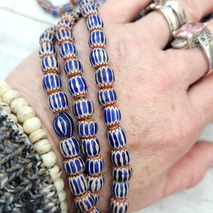 May include: Strands of blue and white striped glass beads with red accents. The beads are oval-shaped and strung together, creating a vintage or antique aesthetic. The beads are held in a hand, suggesting they are jewelry or craft supplies.