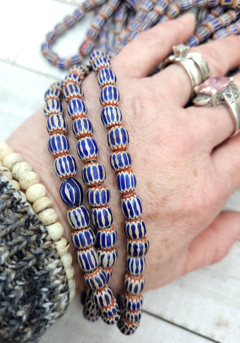May include: Strands of blue and white striped glass beads with red accents. The beads are oval-shaped and strung together, creating a vintage or antique aesthetic. The beads are held in a hand, suggesting they are jewelry or craft supplies.