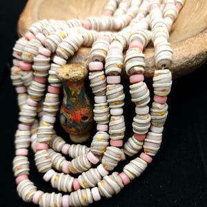 Pode incluir: Um fio de contas de vidro brancas e rosadas com um padrão de espiral. As contas são enfiadas em um cordão e estão dispostas em uma pilha.