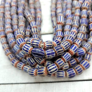 May include: Strands of blue and white striped beads with red accents. The beads have an oval shape and are strung together, creating a layered effect. The beads are displayed on a white wooden surface.