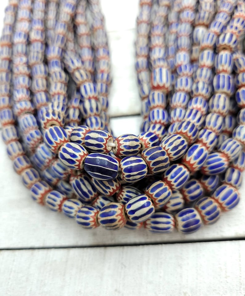 May include: Strands of blue and white striped beads with red accents. The beads have an oval shape and are strung together, creating a layered effect. The beads are displayed on a white wooden surface.