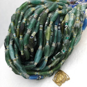 May include: A strand of green, blue, and white glass beads. The beads are various sizes and shapes, some are long and thin, others are round. The beads are arranged in a pile, with some of the beads spilling out of the pile.