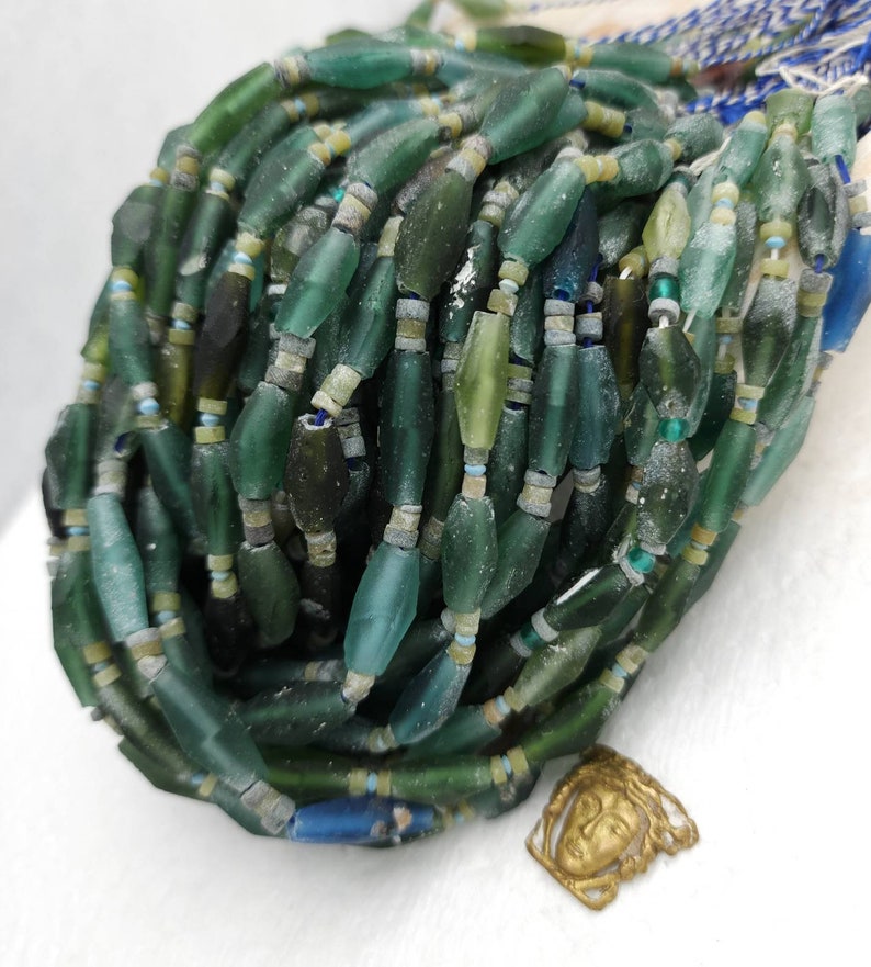 May include: A strand of green, blue, and white glass beads. The beads are various sizes and shapes, some are long and thin, others are round. The beads are arranged in a pile, with some of the beads spilling out of the pile.