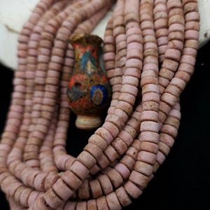 Puede incluir: Múltiples hebras de cuentas cilíndricas rosas. Un pequeño jarrón decorativo con patrones azules, rojos y amarillos se encuentra entre las cuentas. El conjunto se presenta sobre un fondo oscuro. Las cuentas son probablemente de vidrio o cerámica.