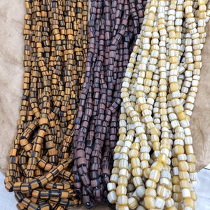 Pode incluir: Três fios de contas de vidro coloridas. As contas são pequenas e cilíndricas com um padrão listrado. Os fios são laranja e preto, marrom e branco, e amarelo e branco.