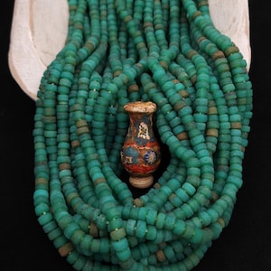 May include: A collection of turquoise-colored, round beads, possibly glass, with a small, decorative vase in the center. The vase is painted with intricate designs in red, blue, and gold. The beads are arranged in a cascading pattern.