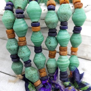 May include: Close-up of a handmade beaded necklace featuring large, seafoam green, diamond-shaped beads. The necklace incorporates smaller, round, dark blue and orange beads. The strands are finished with purple and green fabric tassels, creating a unique, bohemian style.