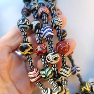 May include: A multi-strand necklace with large, round beads in various colours and patterns. The beads are black, white, red, yellow, blue, and green, with striped and swirled designs. The necklace is strung on black and white cords.