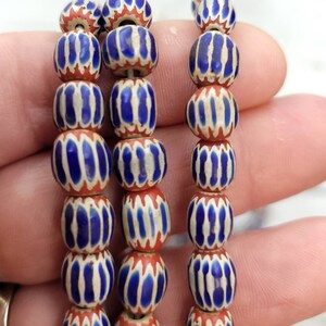 May include: Strands of vintage glass beads. Each bead is a barrel shape, featuring a pattern of blue and white vertical stripes on a red base. The beads are strung together, showcasing their intricate design and color combination.