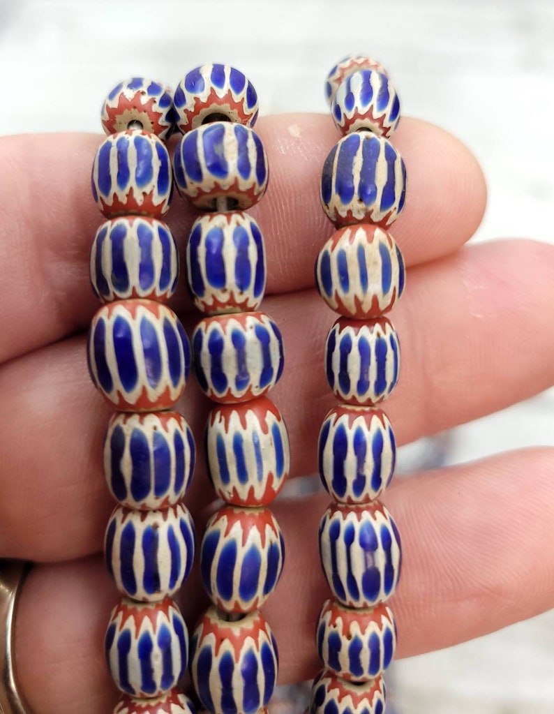 May include: Strands of vintage glass beads. Each bead is a barrel shape, featuring a pattern of blue and white vertical stripes on a red base. The beads are strung together, showcasing their intricate design and color combination.