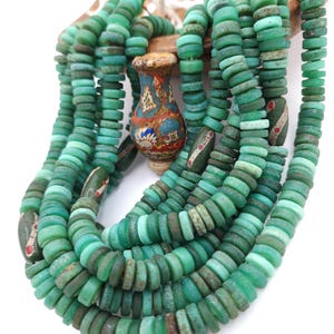 May include: A multi-strand necklace featuring numerous small, circular, matte green beads with a weathered texture. Interspersed among the green beads are several elongated, dark green cylindrical beads with white and red accents, and a central vase-shaped bead.