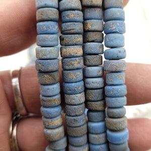 Pode incluir: Um fio de contas de vidro azul com uma superfície áspera e texturizada. As contas são cilíndricas e têm uma forma ligeiramente irregular.