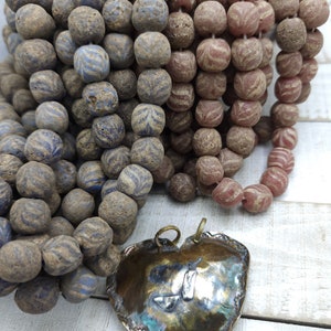 Pode incluir: Duas cordas de contas de vidro, uma azul e outra rosa, com um pingente de coração castanho. As contas têm uma superfície texturizada e salpicada.