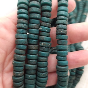 Pode incluir: Um fio de contas de madeira verde-azulada. As contas são redondas e têm uma superfície ligeiramente texturizada.