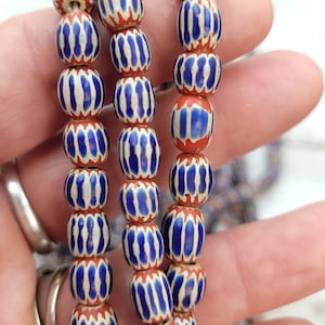 May include: Strands of vintage beads with a repeating pattern. Each bead is a barrel shape with a red base, white stripes, and blue accents. The beads are strung together, creating a decorative element.